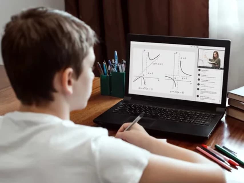 A student following a mathematics lesson on a laptop and taking notes